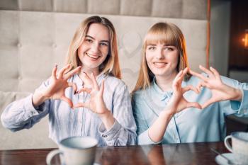 Two smiling girlfriends shows the hearts with fingers, coffee time at the table in cafe. Female friends sitting in restaurant, gossipers meeting