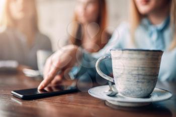 Cup of coffee and mobile phone on wooden table, closeup. Girls leisures in cafe. Female friends sitting in restaurant on couch and drinks coffee, gossipers meeting