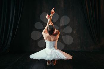 Female classical ballet performer in white dress sitting on the floor, back view. Ballerina training in class with window