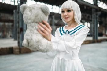 Anime style blonde woman looks at the toy bear. Cosplay girl, japanese culture, doll in dress on abandoned factory