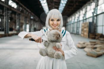 Anime style blonde lady with glass eyes cuts off the head of a teddy bear. Cosplay fashion, asian culture, doll with blade, cute woman with makeup in the factory shop