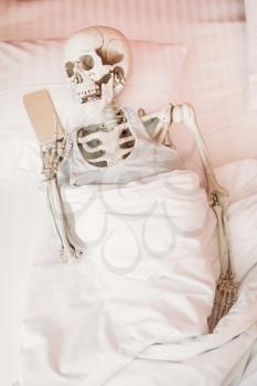 Human skeleton with mobile phone in hand lies in bed, black humor, funny joke