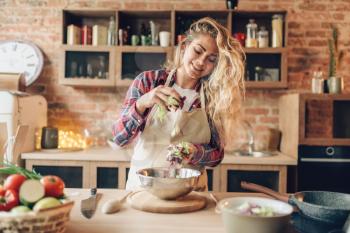 Female cook in apron prepares fresh salad, kitchen interior on background. Housewife making healthy vegetarian food, vegetables preparation