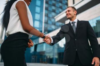 Business woman and businessman shake hands, outdoors meeting of partners, modern office building on background, partnership negotiations. Successful businesspeople