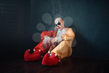 Tired bloody clown with baseball bat sitting on the floor. Man with makeup in carnival costume, crazy maniac