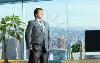 Young businessman in suit stands in the middle of the office, business buildings on background. Businessperson thoughtfully looks into the distance