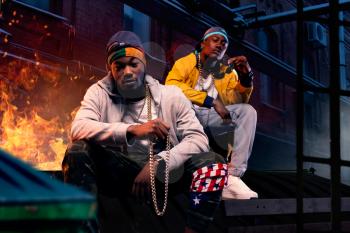 Two black rappers in caps sitting by the fire, night city street on background. Rap performers against cityscape, underground music concert, urban style