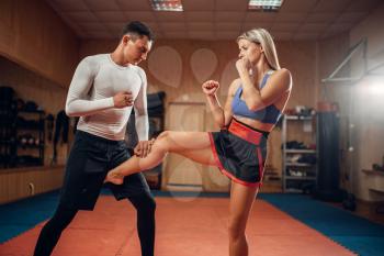 Female person makes a kick in the groin, self defense workout with male personal trainer, gym interior on background. Woman on training, self-defense practice