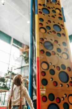 Little girl looks on climbing wall, children game center. Excited childs having fun on playground indoors