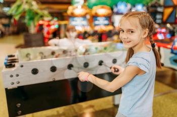 Happy girl at the table soccer in children game center. Smiling female child having fun on playground indoors. Kid plays on football machine in amusement centre