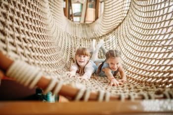 Two funny girls poses in rope net in children game center. Excited childs having fun on playground indoors. Kids playing in amusement centre