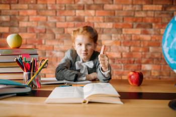 Cute schoolgirl sitting at the table and shows thumbs up. Female pupil at the desk with textbooks, apples and globe