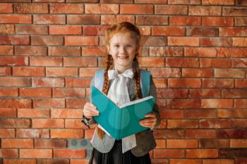 Cute schoolgirl with schoolbag holds an open textbook, brick wall on background. Adorable female pupil with backpack and books poses in the school