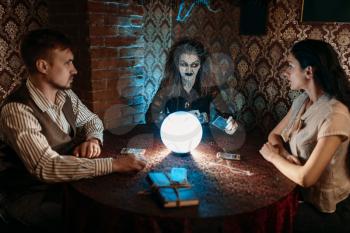 Scary witch reads a magic spell over a crystal ball, young man and woman on spiritual seance. Female foreteller calls the spirits