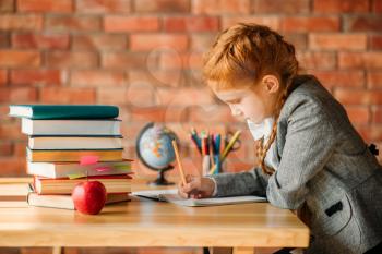 Cute schoolgirl doing homework at the table with textbooks, apples and globe. Female pupil reading a book at the desk