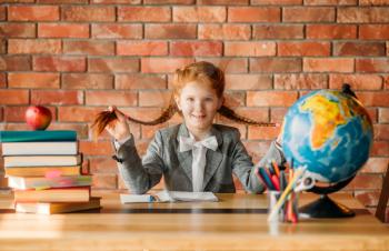 Adorable schoolgirl sitting at the table, front view. Female pupil at the desk with textbooks, apple and globe, young girl doing homework