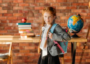 Cute schoolgirl with schoolbag, side view. Female pupil with backpack, desk with textbooks, apple and globe on backgrpund, young girl in the school