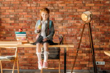 Cute schoolgirl sitting on the table, front view. Female pupil on the desk with stack of textbooks and apple on the top