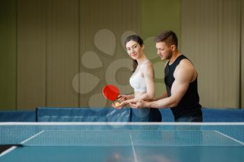 Man teaches a woman to play ping pong, training indoors. Couple in sportswear holds rackets and plays table tennis in gym