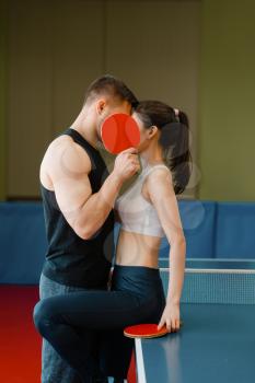 Couple with rackets kissing at the ping pong table with net indoors. Man and woman in sportswear, table-tennis training game in gym