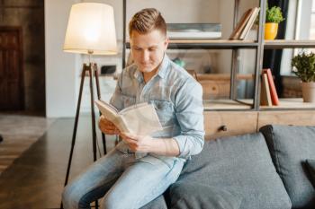 Young man sitting in couch and reading a book. Male person reads in living room, floor lamp and shelf on background