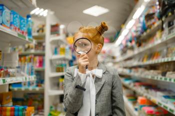 Little school girl looks through magnifying glass, shopping in stationery store
