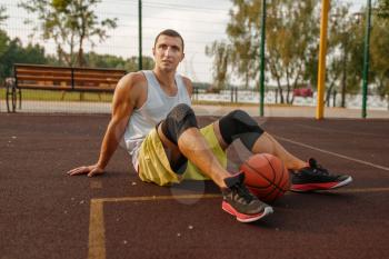 Basketball player with ball sitting on the ground on outdoor court. Male athlete in sportswear resting after streetball training