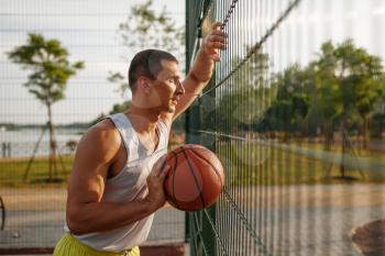 Male basketball player with ball standing at the mesh fence on outdoor court, side view. Male athlete in sportswear on streetball training, summer stadium