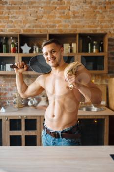 Muscular man with naked body holds frying pan on the kitchen. Nude male person preparing breakfast at home, food preparation without clothes