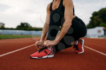 Female runner in sportswear tying her shoelaces, training on stadium. Woman doing stretching exercise before running on outdoor arena