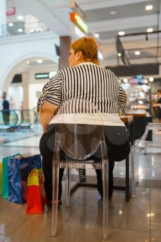 Fat woman sitting at the table in fast food restaurant, back view. Overweight female person in mall fastfood, obesity problem