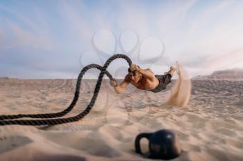 Male athlete with battle ropes, shoot in jump, flying sand effect, desert at sunny day. Strong motivation in sport, strength outdoor training