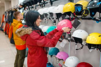Couple at the showcase choosing helmets for ski or snowboarding, side view, shopping in sports shop. Winter season extreme lifestyle, active leisure store, customers buying protect equipment