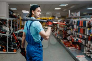 Male worker in uniform holds two axes in tool store. Choice of professional equipment in hardware shop, instrument supermarket