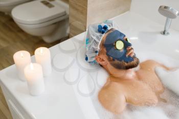 Man applies face mask and relax in bath with foam, morning hygiene. Male person resting in bathroom, skin and body treatment