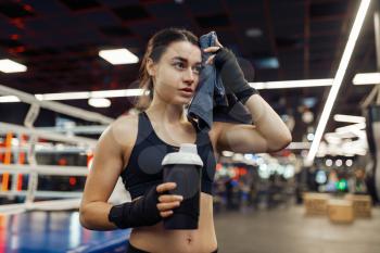 Tired woman wipes her sweat after boxing training, ring on background. Female boxer in gym, girl kickboxer in sport club, kickboxing workout