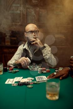 Bearded poker player with cigar, casino. Games of chance addiction. Man leisures in gambling house. Cards, chips and whiskey on gaming table with green cloth