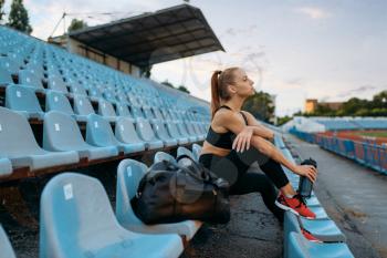 Female runner in sportswear sitting on tribune and drinks water, training on stadium. Woman doing stretching exercise before running on outdoor arena