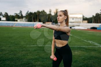 Female runner in sportswear holds dumbbells, workout on stadium. Woman doing stretching exercise before running on outdoor arena