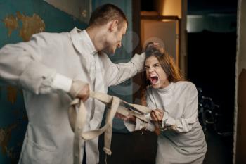 Male psychiatrist and crazy female patient in a fit of rage, mental hospital. Woman in strait jacket undergoing treatment in clinic for the mentally ill