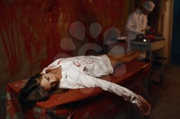 Bloodied body of young female patient, crazy male psychiatrist on background, mental hospital basement. Victim of doctor maniac lying on surgical table in clinic for the mentally ill