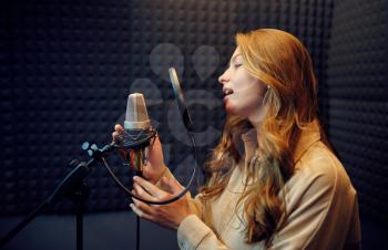 Female singer in headphones sings a song at micriphone, recording studio interior on background. Professional voice record, musician workplace, creative process, modern audio technology