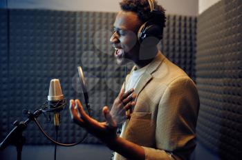 Male singer in headphones sings a song at micriphone, recording studio interior on background. Professional voice record, musician workplace, creative process, modern audio technology