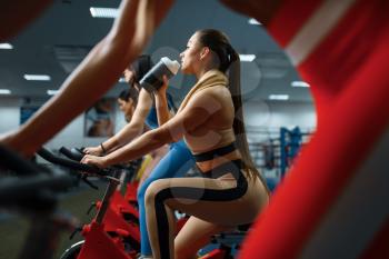 Woman drinks water on a stationary bike in gym. People on fitness workout in sport club, athletic girls in sportswear on training indoors