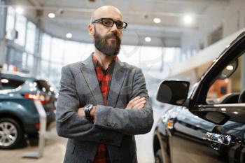 Bearded man in glasses poses at the pickup truck in car dealership. Customer in vehicle showroom, male person buying transport, auto dealer business