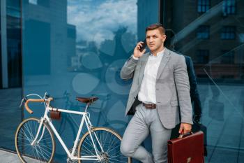 Businessman in suit talking by mobile phone at the bicycle in downtown. Business person riding on eco transport on city street