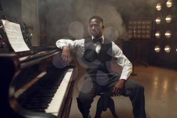 Ebony grand piano player, jazz performer on the stage with spotlights on background. Negro musician poses at musical instrument before concert