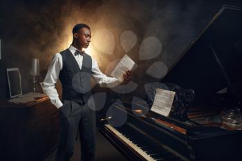Ebony pianist with music notebook in his hands on the scene with spotlights on background. Negro performer poses at musical instrument before concert