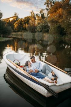 Love couple lying in a boat on quiet lake at summer day at sunset. Romantic meeting, boating trip, man and woman walking along the river