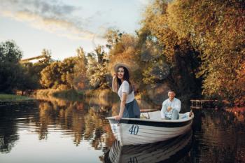 Pretty lady poses in boat on quiet lake at summer day. Romantic date, boating trip, man and woman walking along the river